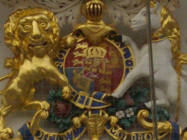 The image depicts an ornate crest featuring a gold lion and a white unicorn on either side of a shield adorned with a colorful heraldic design. The crest is surrounded by a golden ribbon with inscriptions. The overall image suggests that it might be part of a coat of arms or a ceremonial decoration, and it is presented with a slightly blurred effect, indicating that the photo might have been taken with a camera with a limited resolution, such as an older model like the Pentax Optio E30.