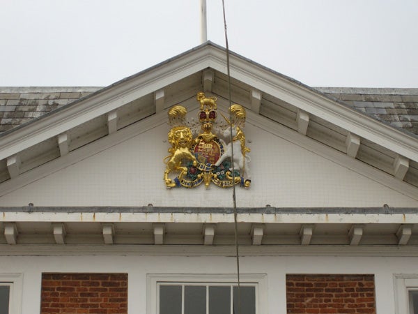 The image displays a golden royal crest with intricate designs mounted on the white pediment of a brick building. The photograph is taken during the day under overcast conditions, capturing fine details of the emblem and the architectural elements of the building.
