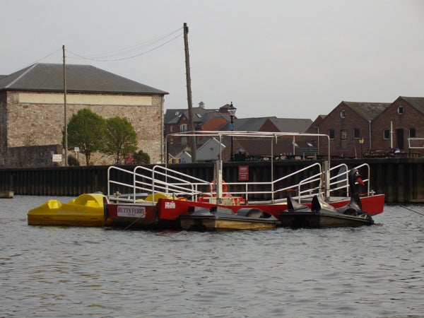 Photograph depicting a small yellow pedal boat and a larger red and white ferry boat floating on calm water with historic buildings in the background, illustrating the color reproduction and clarity of a photo taken by the Pentax Optio E30 digital camera.