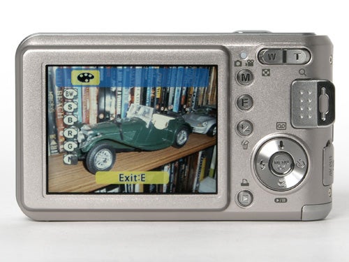 Samsung L700 digital camera with a photo displayed on its LCD screen showing a vintage car toy on a wooden floor with bookshelves in the background.