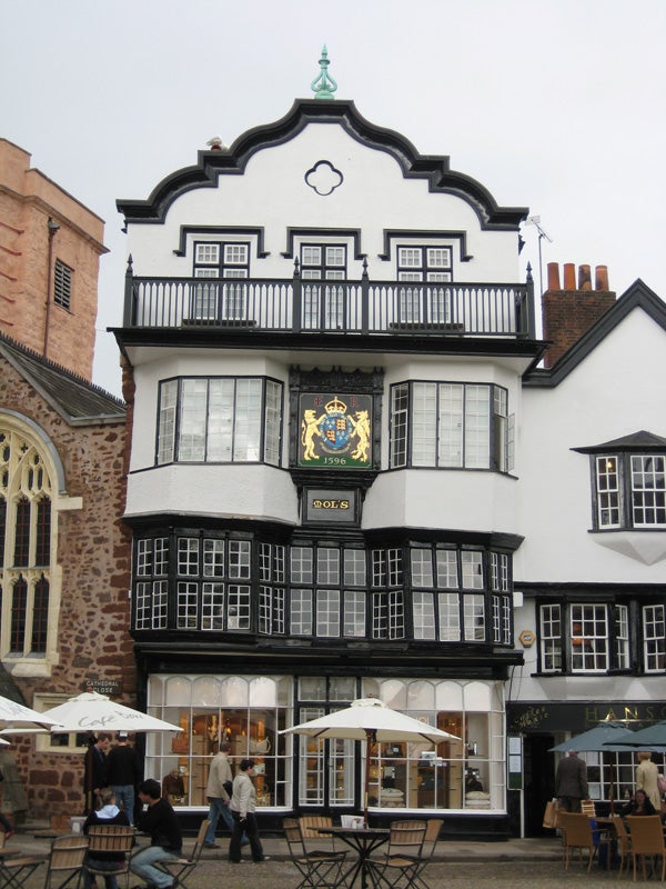 A charming traditional multi-storied building with a detailed façade, featuring black and white Tudor-style exteriors with a large coat of arms sign dated 1596, flanked by a modern café setting with patrons and umbrellas in the foreground.