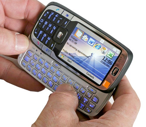 A person holding an Orange SPV E650 smartphone with a slide-out QWERTY keyboard and the screen displaying the Windows Mobile interface with icons for calendar, mail, and other apps.