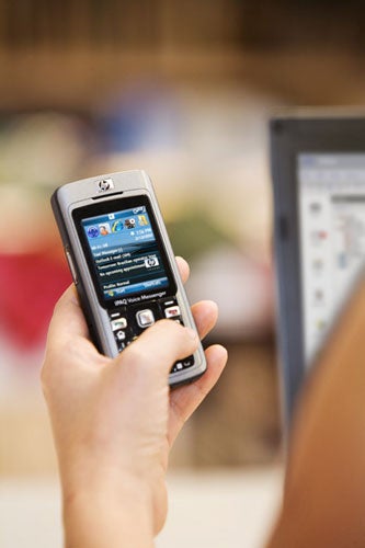 A person holding a Hewlett Packard iPAQ 514 Voice Messenger with the screen display visible, showing the start menu with applications, in focus against a blurred background of what appears to be a computer screen.