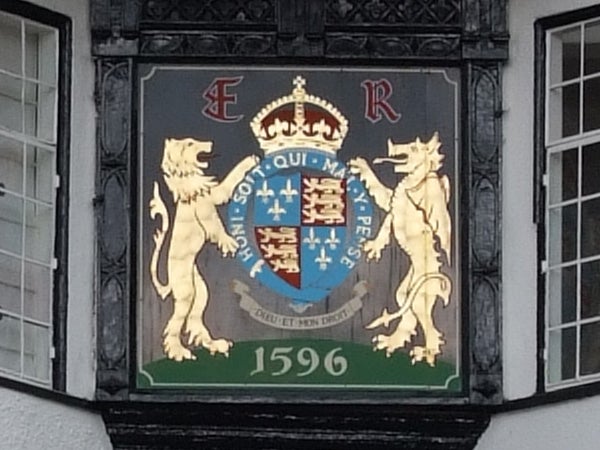 The image shows a detailed royal crest with two golden lions, flanking a shield adorned with various heraldic symbols, against a black and white timber-framed building. The crest is topped with a crown and has the initials 'E R' on either side, with a Latin motto below. The date 1596 is prominently displayed at the bottom against a green background.