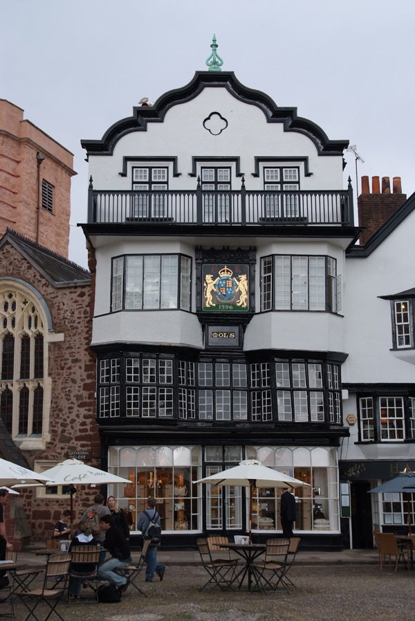 A traditional half-timbered building with a white facade, black wooden beams, and a distinct coat of arms dated 1596. The building features a balcony on the upper floor and is located next to a cafe with outdoor seating and patrons.