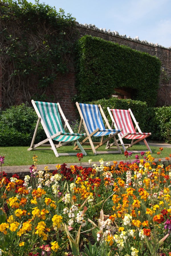 Three striped deck chairs in green, blue, and red set against a colorful flowerbed and a brick wall with green ivy.