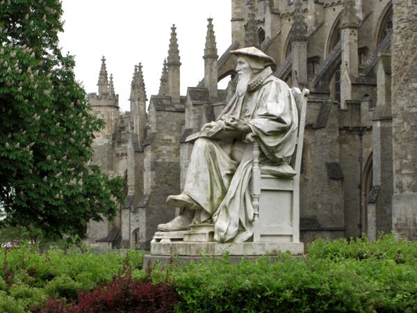 Statue of a seated bearded figure with a contemplative expression in front of a Gothic cathedral surrounded by greenery.