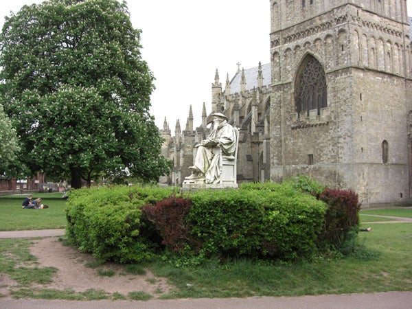 A clear daytime photo featuring a historical stone statue in front of an old cathedral surrounded by greenery, with people lounging on the grass in the background, showcasing the imaging capabilities of the Casio Exilim EX-Z1050 digital camera.