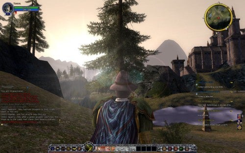 Screenshot from the video game Lord of the Rings Online: Shadows of Angmar showing a player's character dressed in a cloak and wizard's hat standing in a serene landscape with trees, a lake, distant mountains, and an in-game chat window visible at the bottom left corner. The game's user interface includes a mini-map, character status bars, and ability icons along the screen's bottom edge.