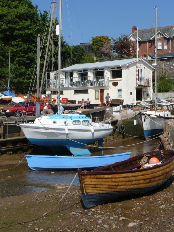 This image shows a clear, sunny day at a boatyard with multiple boats moored in a low tide. In the foreground, there's a wooden rowboat with an orange buoy inside, and its color appears faded but rich in historical character. The middle ground presents two larger boats; the nearest is a blue sailboat with its sail down, securely tied to the dock. Behind the boats, there's a gathering of people enjoying the weather on a balcony of a white building with red trim, which is likely a part of the boatyard facilities or a nearby café. The overall scene suggests a leisurely, relaxed atmosphere in a maritime setting.
