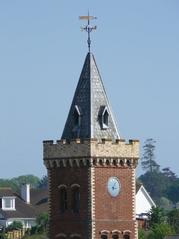 A clear and focused image showing the top section of a brick tower with a pointed roof, a weathervane on top and a blue clock face visible on the tower. The background is a clear blue sky with a hint of green foliage at the bottom.