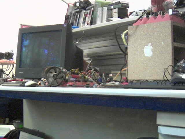 Image displaying a cluttered desk with various electronic components and tools, a computer monitor, a cardboard box with an Apple sticker, and a disassembled device which may represent a DIY or repair setting associated with a Trendnet TV-IP400W review.