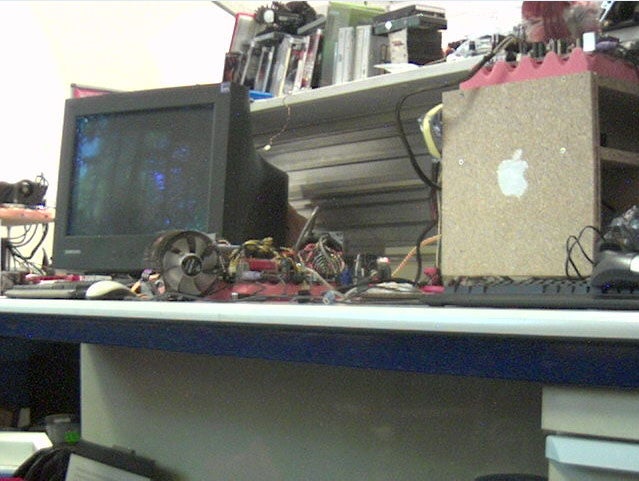 Cluttered workspace with various electronic components and a CRT monitor, likely depicting a tech enthusiast's or hobbyist's desk.