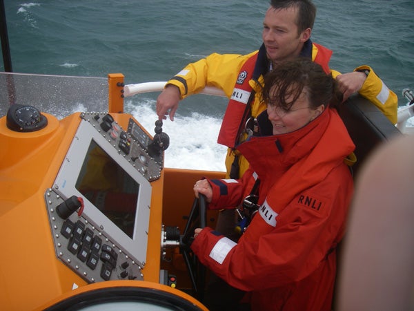 Two individuals in bright life jackets piloting a boat with modern navigation equipment under overcast weather conditions.