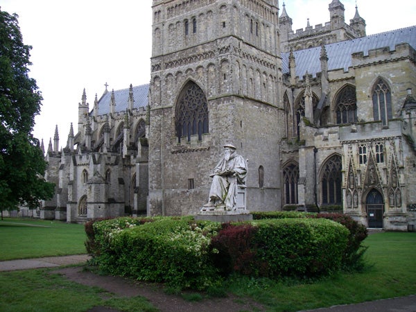 This is a photograph of a historic cathedral with intricate Gothic architecture, featuring a statue in the foreground nestled within a well-manicured hedge. The image is captured under an overcast sky which allows for even lighting on the scene.