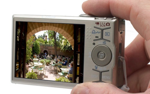 Hand holding Canon IXUS 90 IS camera displaying a courtyard scene.Hand holding Canon IXUS 90 IS camera displaying image on screen.