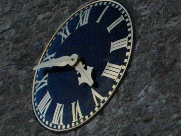Low-light photo of a wall-mounted clock taken with Olympus E-3.Oval clock showing time on stone wall at dusk.