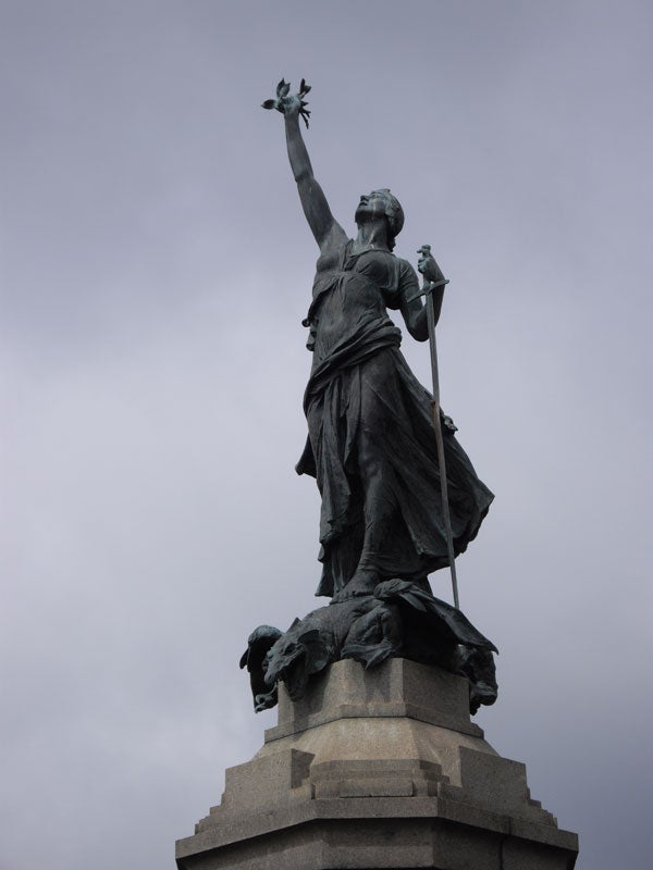Statue against cloudy sky background.Bronze statue of a figure holding a raised torch and a spear.