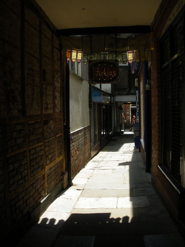Narrow alley with sunlight and shadows, possibly taken with Pentax camera.Narrow alleyway with sunlight and shadows captured by camera.