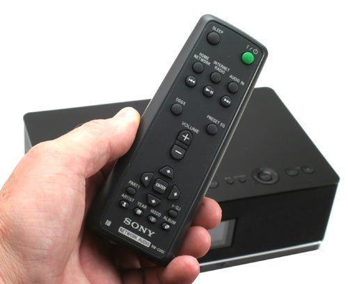 Hand holding Sony Giga Juke remote with speaker in background.Hand holding Sony Giga Juke remote with system in background.