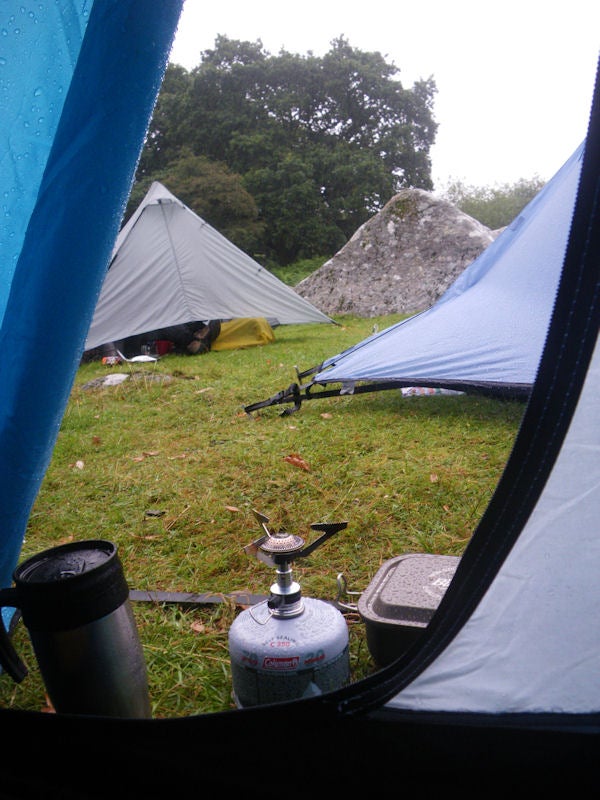 View from inside a tent at a rainy campsite with gear.View from inside a tent at a campsite with outdoor stove.