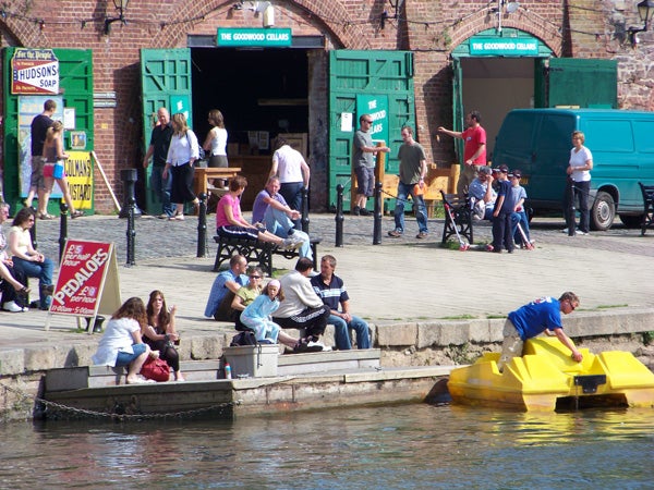 People by the water near pedal boats and market stalls.People enjoying a sunny day by the waterfront.