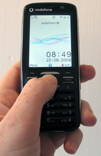 Hand holding a Vodafone 725 mobile phone displaying the time and date.