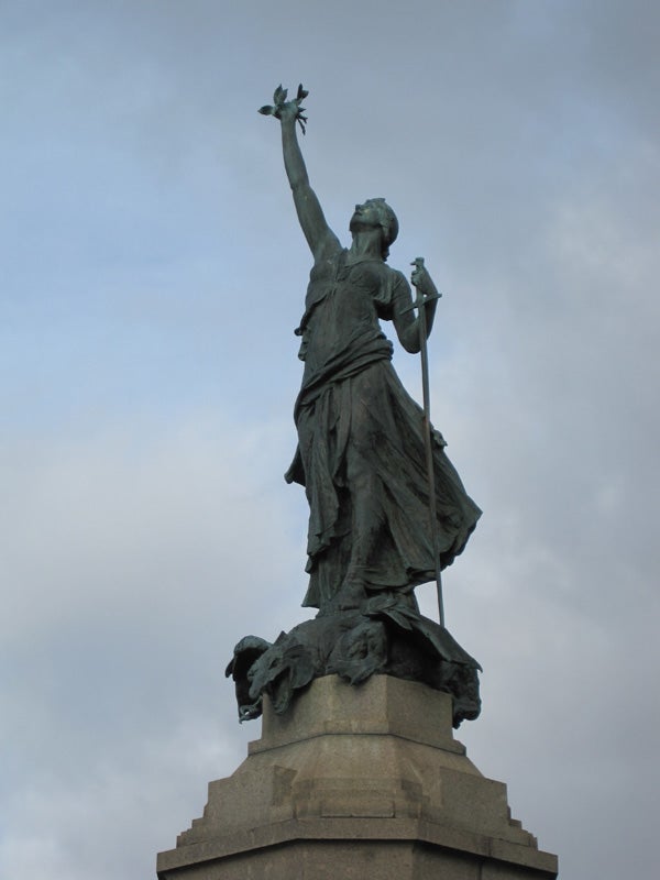 Bronze statue of a figure holding a torch and spear against sky