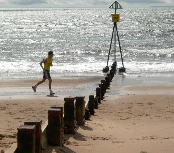 Person jogging on beach beside wooden posts and navigational marker.
