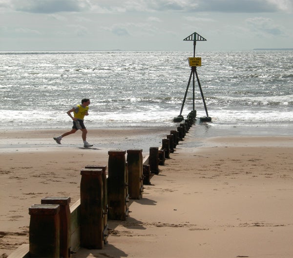 Person jogging on beach near warning sign and groynes.