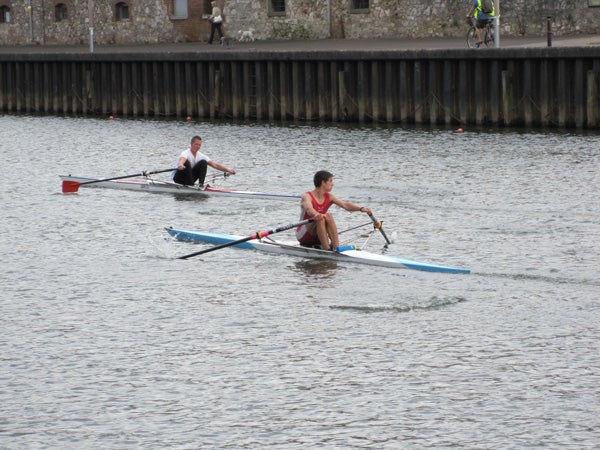 Two people rowing on calm water with urban background.