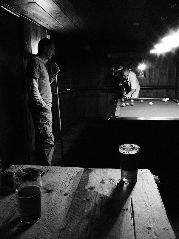 Black and white photo of two men playing pool with a beer on table.