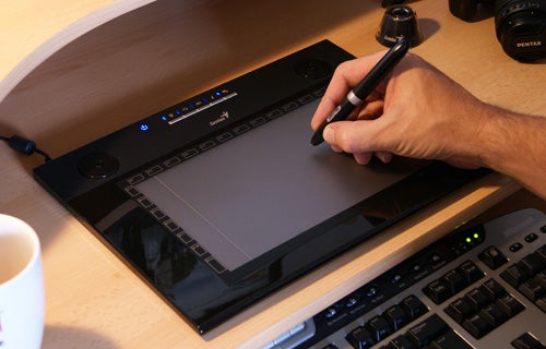 Hand using Genius G-Pen M609X pen tablet next to keyboard.