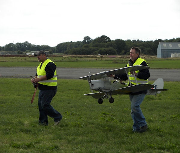 Two men carrying a model airplane across a grass field.