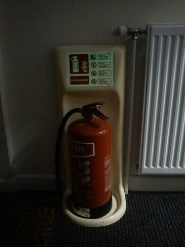 Fire extinguisher standing by a white wall near a radiator.