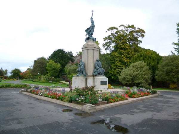 Statue in a park with flowers and trees, under cloudy sky.