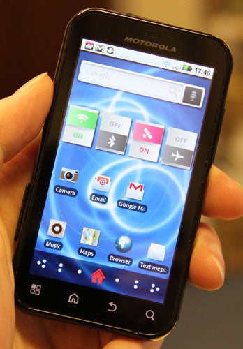 Hand holding a Motorola Defy smartphone displaying the home screen.