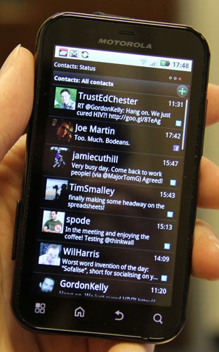 Hand holding a Motorola Defy displaying contacts and tweets.