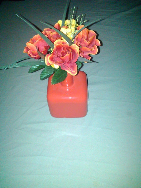 Artificial orange roses in a red square vase.
