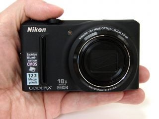 Hand holding Nikon Coolpix S9100 camera with specifications.