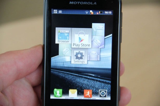 Hand holding Motorola Defy Mini displaying homescreen icons.