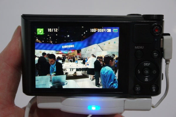 Hand holding Samsung WB150F camera with trade show on display screen.
