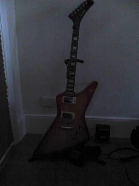 Dark image of an electric guitar standing against a wall.
