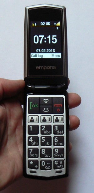 Hand holding an open flip phone displaying time and menu options.