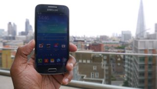 Hand holding Samsung Galaxy S4 Mini with cityscape background.