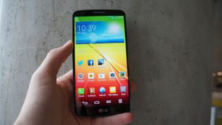 Hand holding an LG G2 smartphone displaying home screen.