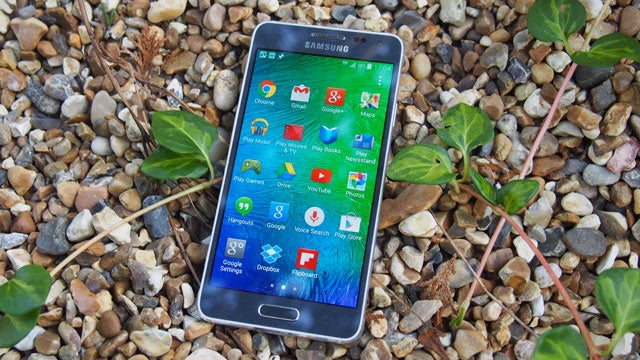 Samsung Galaxy Alpha smartphone lying on pebbles with screen visible.
