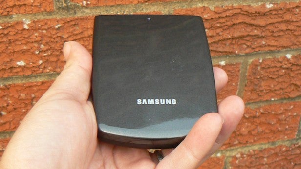 Samsung UHD Video PackHand holding a compact Samsung electronic device against a brick wall.