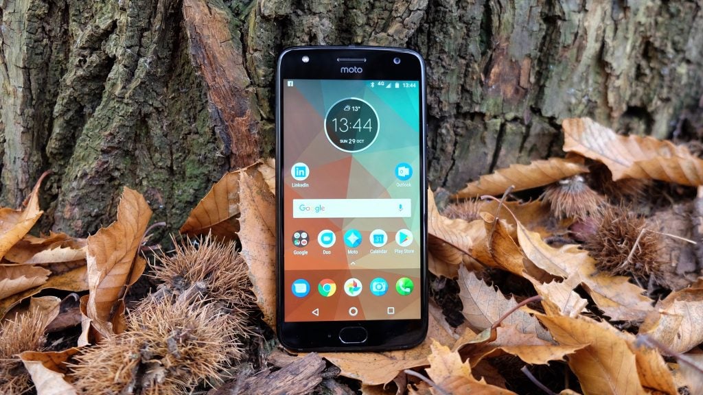 Moto X4 smartphone on forest floor surrounded by autumn leaves.Hand holding Moto X4 displaying features on screen.