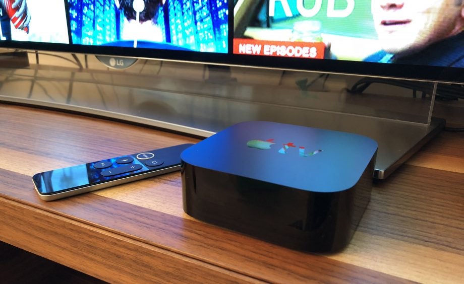 Apple TV 4K with remote on wooden surface, TV in background.